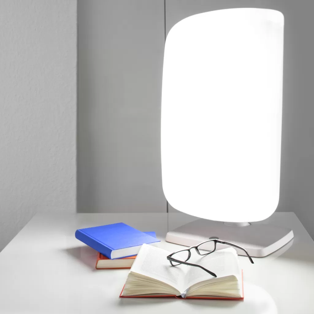 Light box therapy lamp on a desk with a book and glasses, offering seasonal affective disorder (SAD) support during winter.