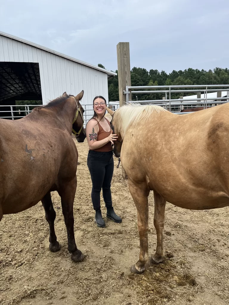 Woman standing with two horses, smiling as the horses nuzzle close to her, seeking affection.