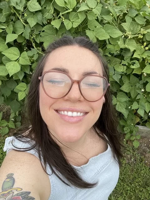 Woman standing in a lush garden with green beans growing behind her, smiling with her face lifted toward the sun.