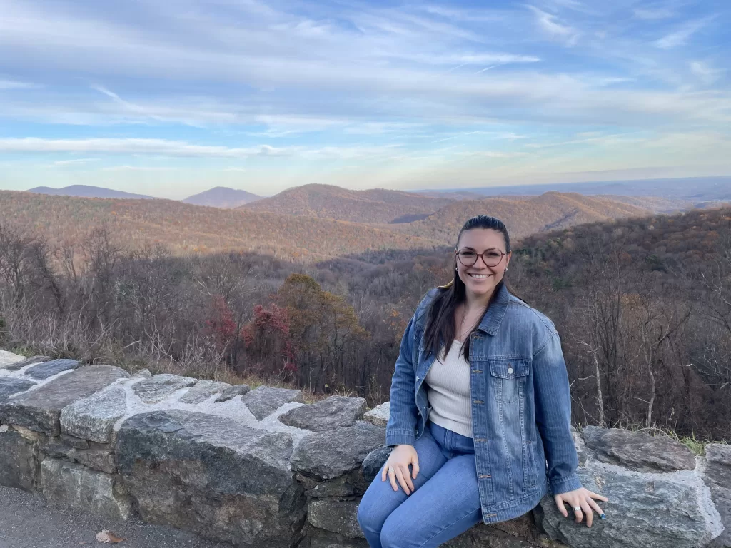 Woman sitting on a short rock wall, smiling at the camera with the mountains of Luray, Virginia, in the background.