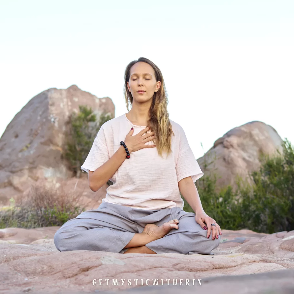 Woman meditating in Sukhasana with hand on heart, eyes closed, in a peaceful desert setting. Yoga for autoimmune relief and stress reduction.