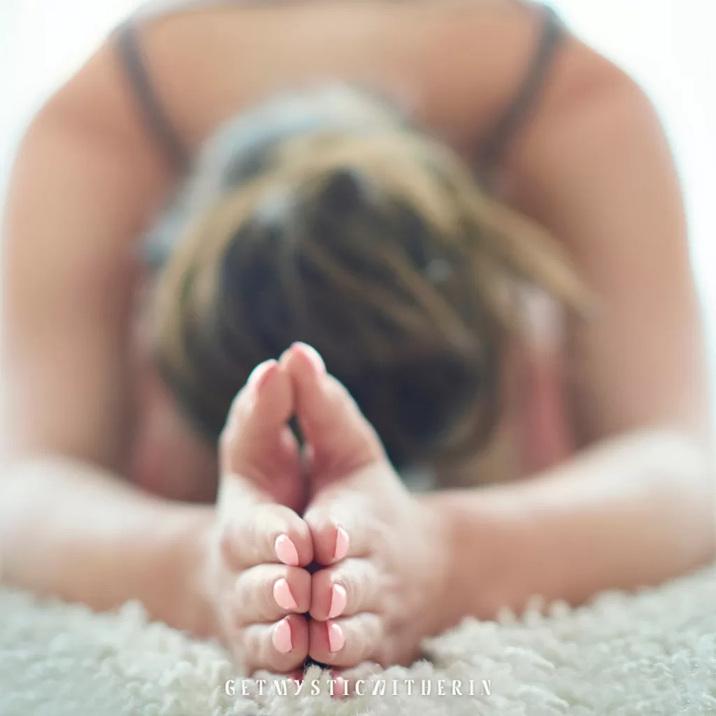 Close-up of hands in Anjali Mudra (prayer position) resting on a yoga mat, with blurred figure in Child’s Pose. Close-up of hands in Anjali Mudra (prayer position) resting on a yoga mat. Restorative yoga for autoimmune relief and relaxation.